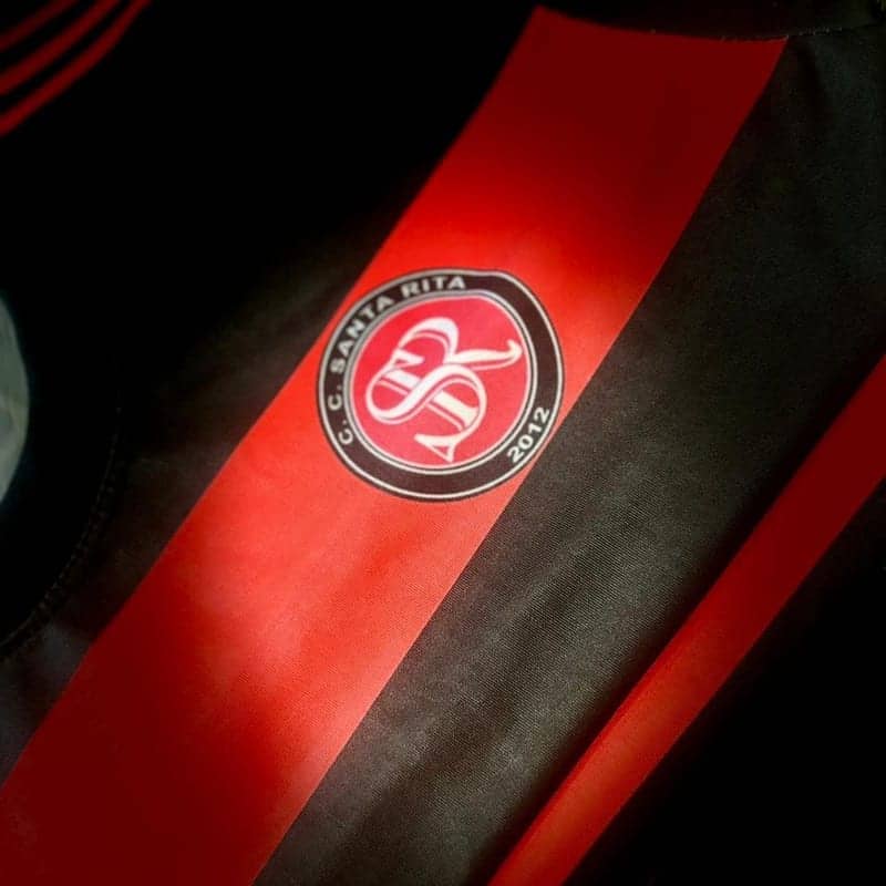 Primer plano en diagonal de una camiseta negra con una banda roja ancha: la luz cruzada deja ver la trama del tejido, y sobre la banda va el escudo circular de una institución en rojo, blanco y negro.