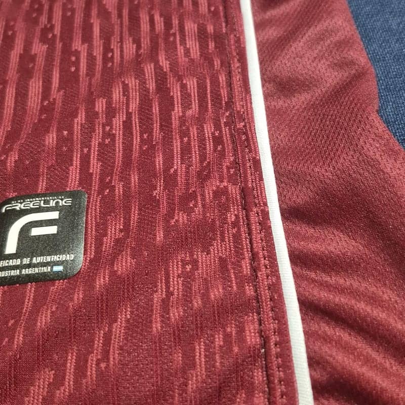 Primer plano del costado de una camiseta bordó: se ve la trama en relieve del tejido, el vivo blanco que corre por la costura lateral, y la etiqueta de autenticidad de Free Line cosida al ruedo.