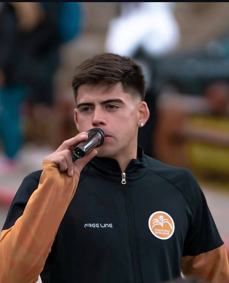 Un joven habla por un micrófono de mano con una campera de media cremallera negra y naranja de la marca, con el escudo de una institución en el pecho.
